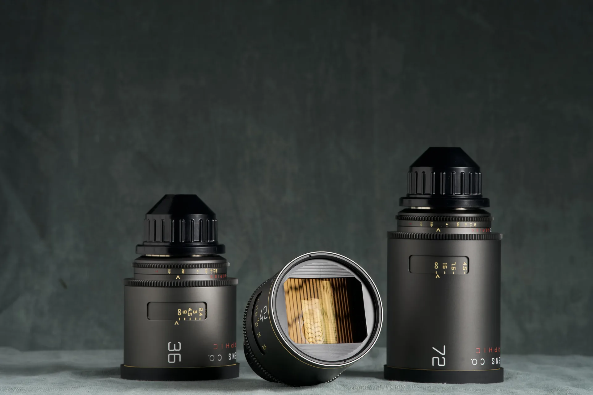 Three Atlas Mercury anamorphic cine lenses on a pale cloth against a green-grey painted backdrop, the centre lens turned to show its anamorphic front element.